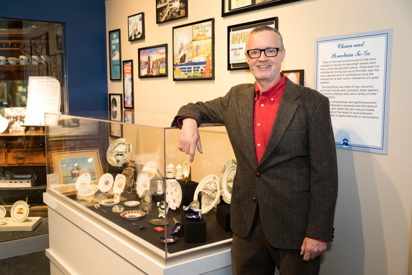 Man in glasses standing by museum display case with collectibles; wearing a red shirt and blazer.
