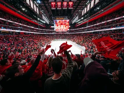 By Allison Farrand. Fans in a stadium cheering a goal, waving red towels, ice rink in the center, and a large screen displaying "Goal!"