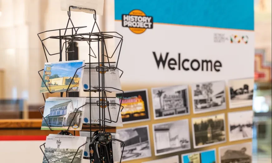 Postcard rack in front of a "Welcome" exhibit sign with historical photos.
