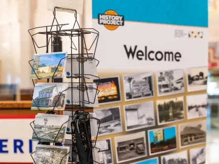 Postcard rack in front of a "Welcome" exhibit sign with historical photos.