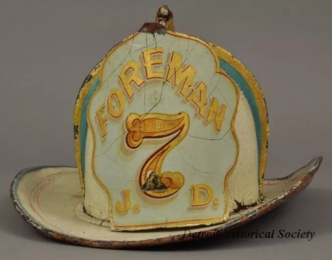 White leather "leatherhead" style firefighter's helmet with red, blue, and gold trim. Front crest reads "Foreman" in ornate curving letters above the number 7. The initials J.D., for its wearer Jeremiah Dwyer, are at the crest's bottom corners. This front crest is supported by the beak and neck of a gold eagle, extending from the helmet's peak.