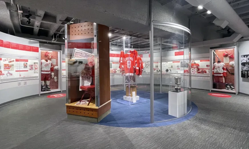 Hockey museum exhibit with red jerseys displayed in glass cases, trophies, and historical photos on walls.