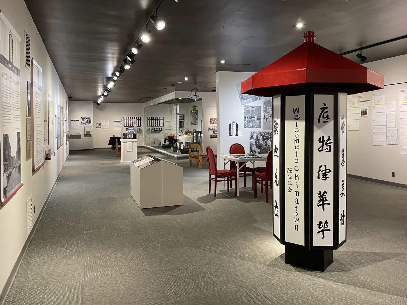 Museum exhibit with historical photographs and displays, featuring a lantern-like column with red roof and Chinese characters.