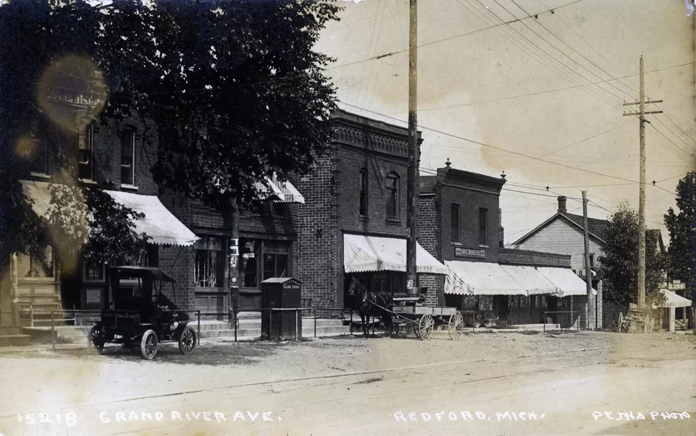 Historic street scene with vintage buildings, cars, and horse-drawn wagon under an overcast sky.