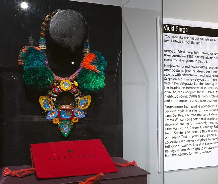 Colorful beaded necklace with pom-poms displayed in a glass case alongside a red envelope and text panel.